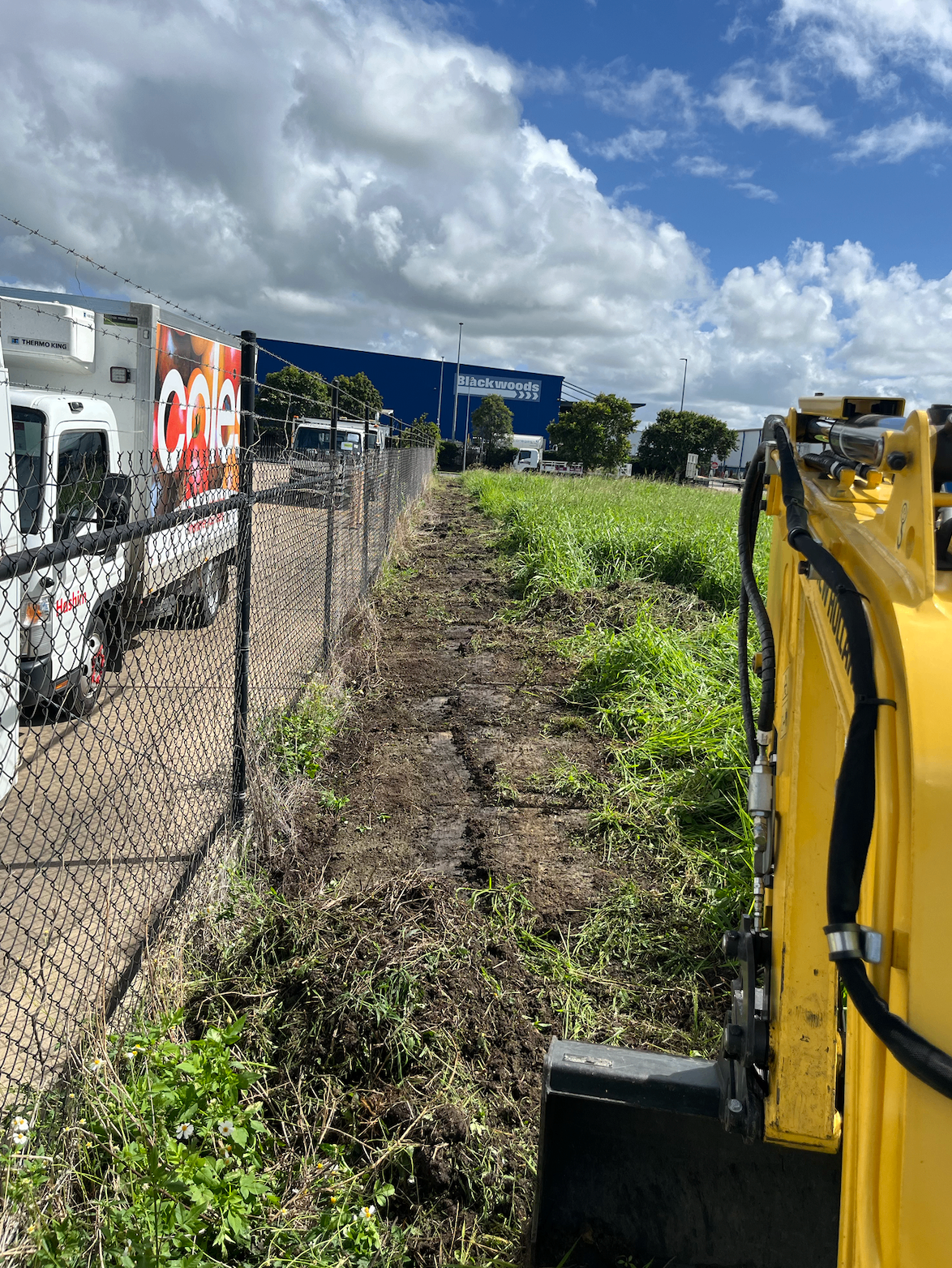 Excavator trenching along a fence line at a commercial site